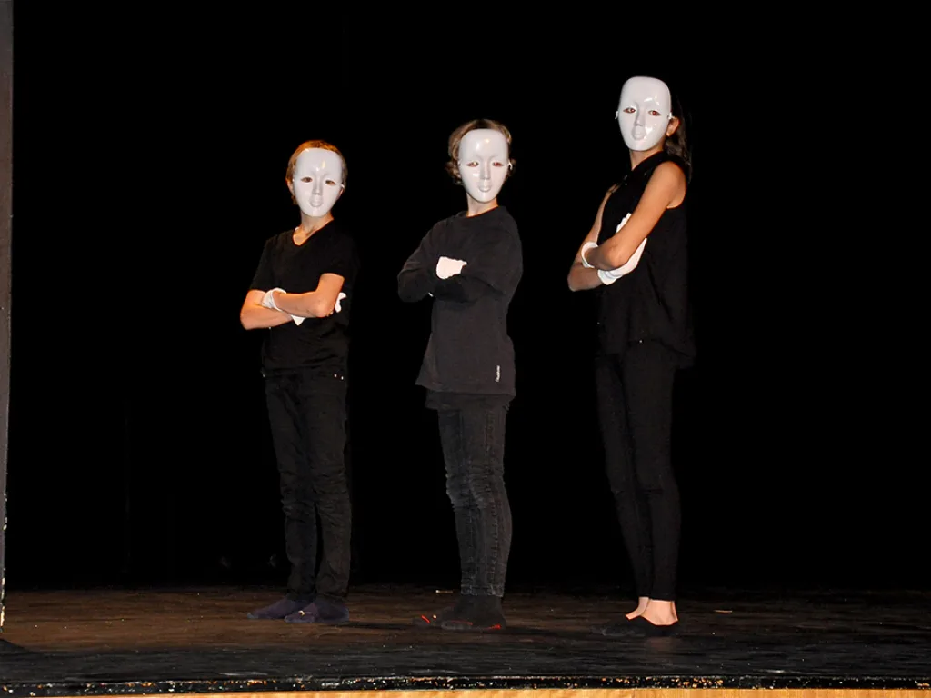 Drei Darsteller mit weißen Masken stehen im Dunkeln auf der Bühne.